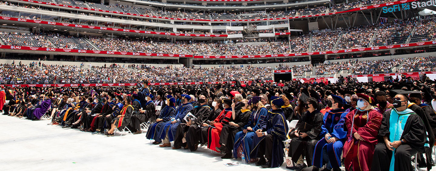 A sweeping view from field level of all graduates with thousands of guests in the the seating above field level