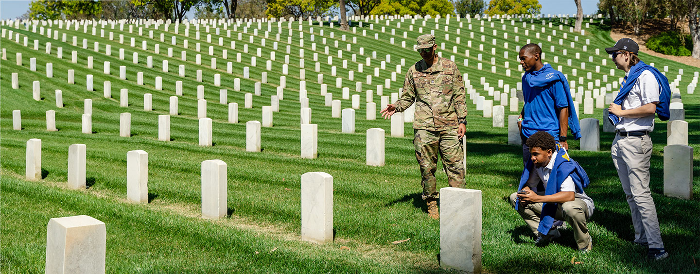 Two high school students speaking with a uniformed soldier while looking at grave stones in a large cemetary